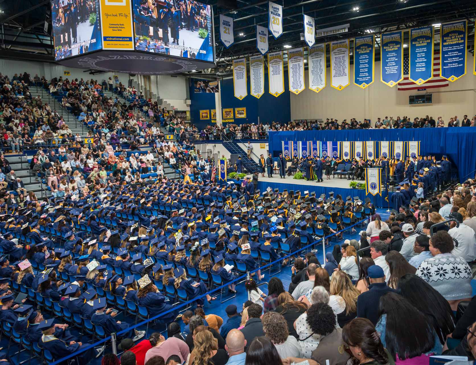 High-angle wide shot of a crowded graduation ceremony inside the VU P.E. Complex. Graduates in blue caps and gowns sit on the floor facing a stage with banners above and audience in bleachers.