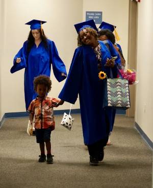Two ABE graduates walk down a hallway, one is holding hands with her child while carrying the other child.