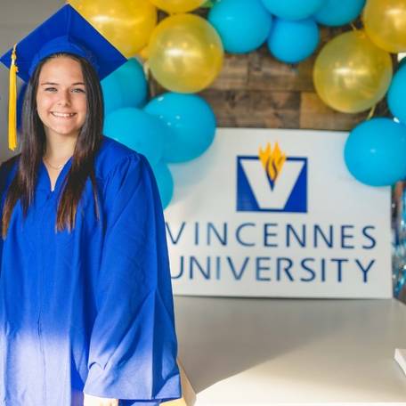 A female graduate stands for a picture near a Vincennes University sign with gold and blue balloons around the sign