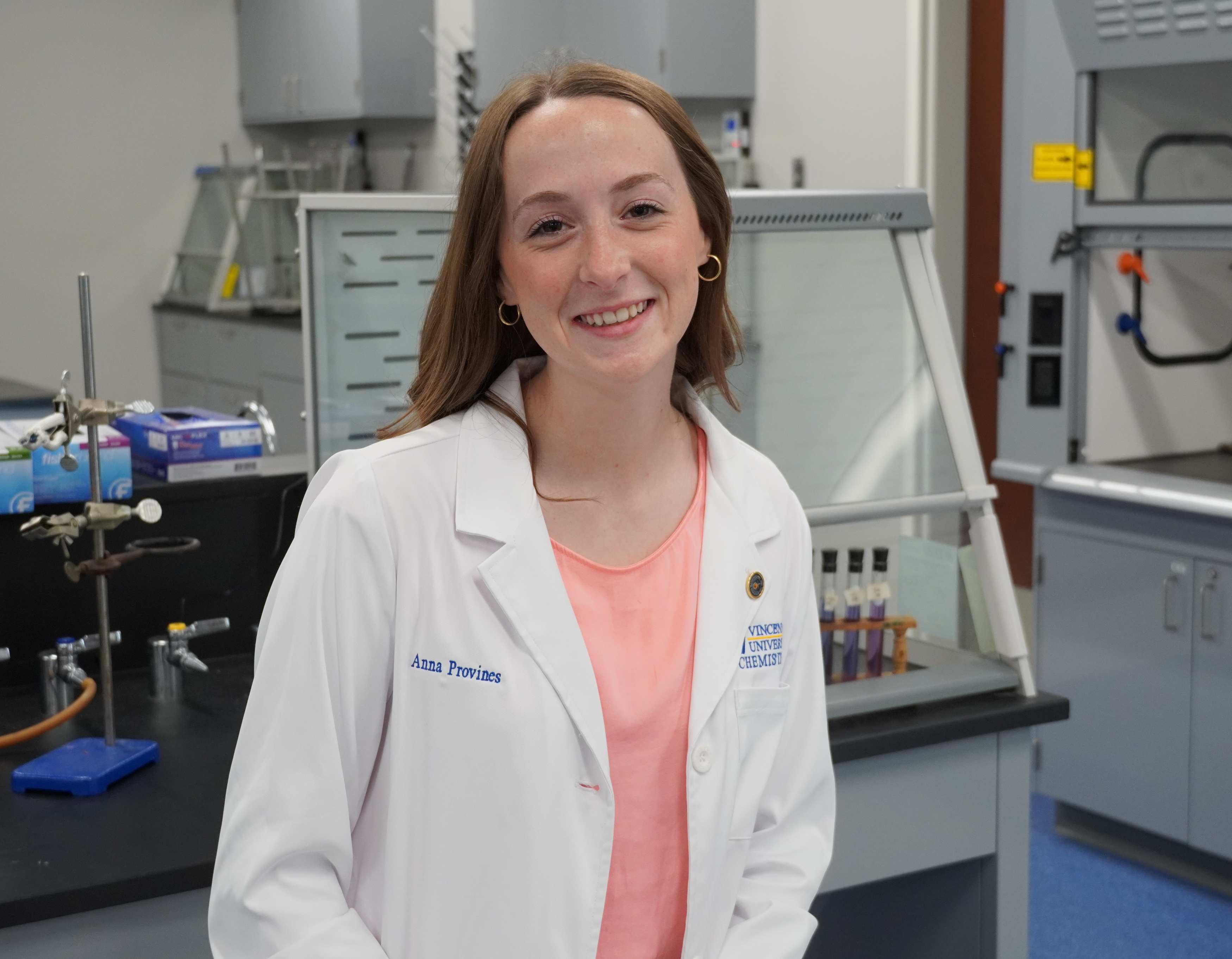  A smiling Anna Provines in a white lab coat embroidered with her full name and a university logo sits in a chemistry lab with beakers and equipment.