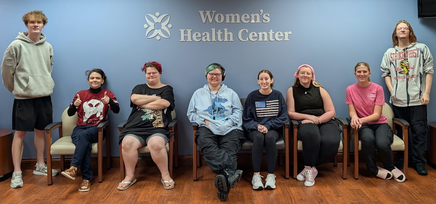 A group photo of eight college students seated and standing in front of a blue wall with a sign that reads 
