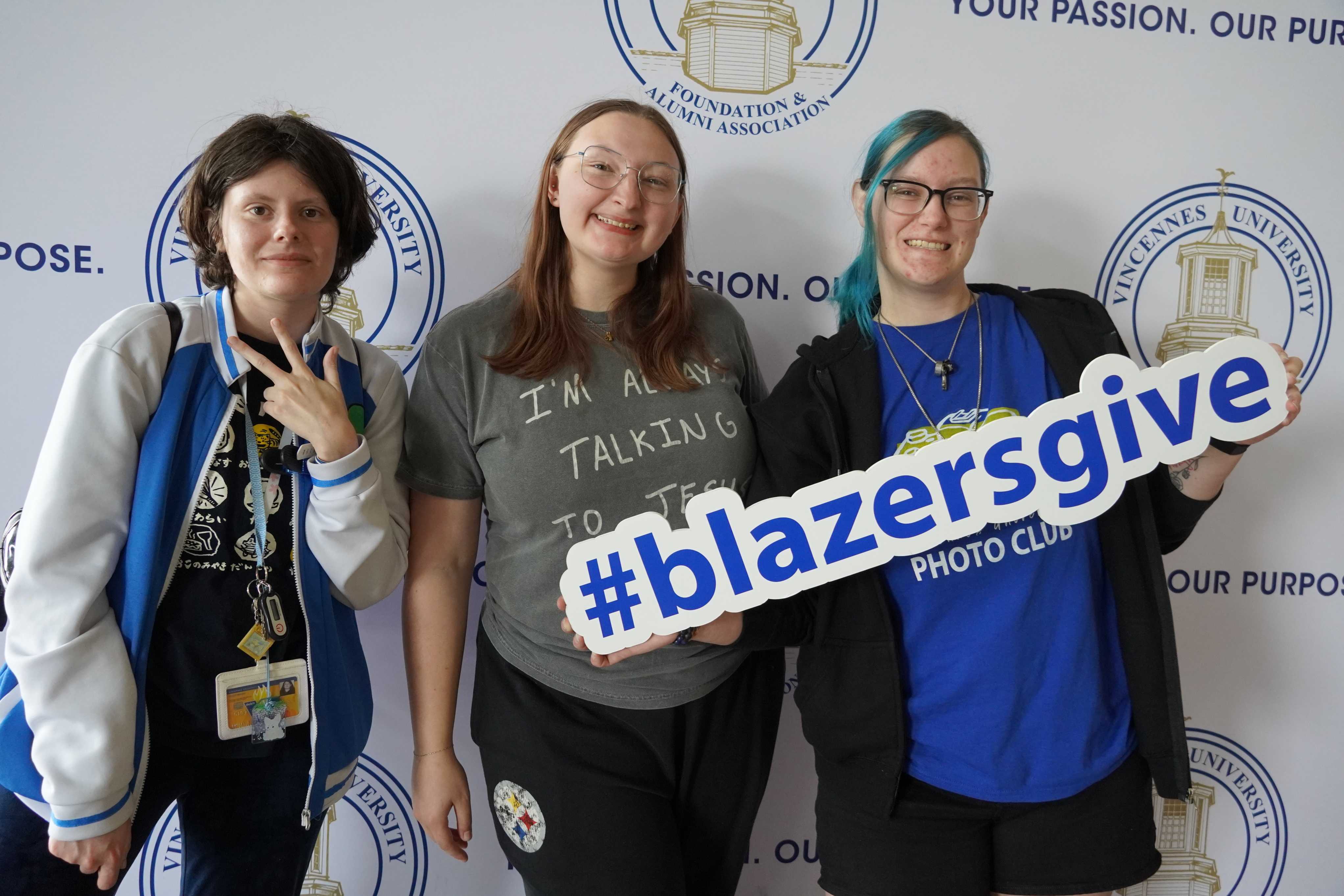 Three students smiling and holding a Blazers Give sign.