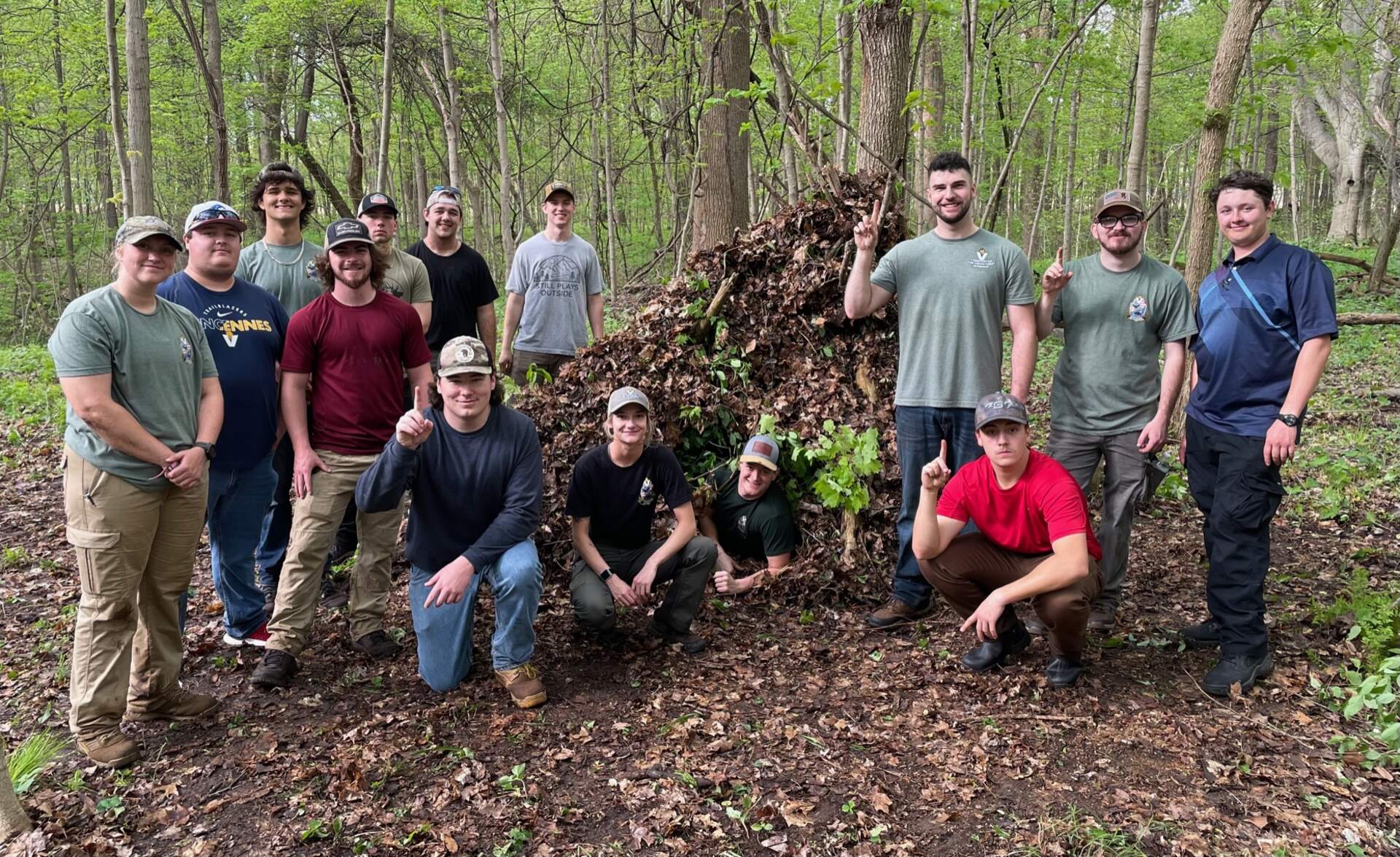 A group of about a dozen young adults stand outside in front of a large pile of dead leaves and sticks.