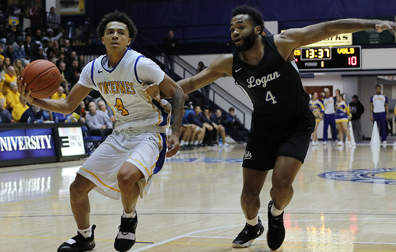 Two basketball players in action on a court; one in a white uniform dribbling the ball while the other in black defends closely. Audience in the background.