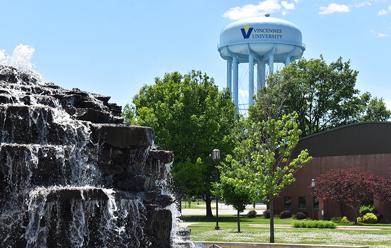 A cascading stone waterfall flows on the left, with lush green trees and a red brick building in the background. A water tower marked "Vincennes University" rises above, set against a clear blue sky. The scene conveys tranquility and academia.