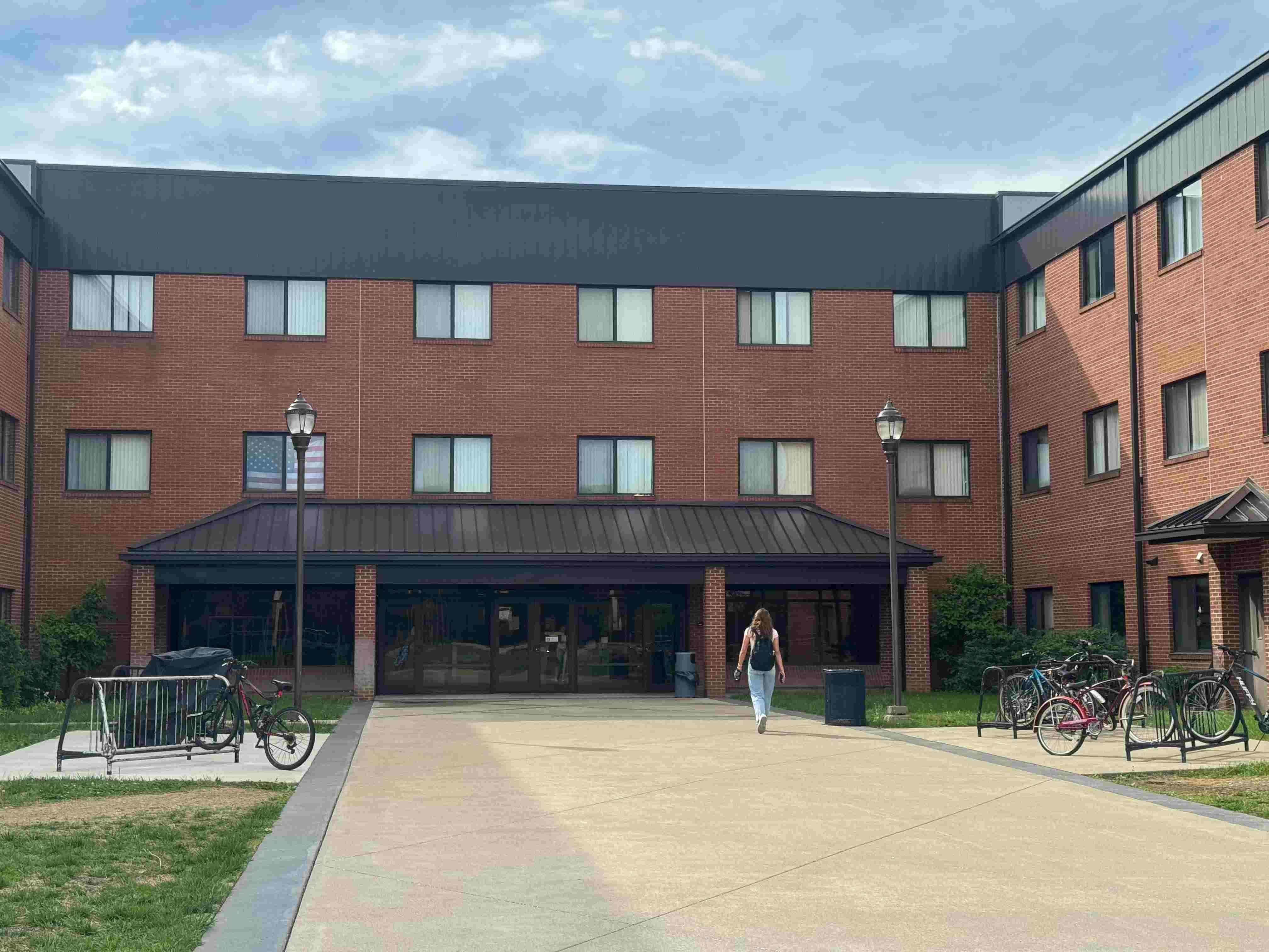 Brick residence hall building with a female college student walking toward the glass entrance. Bicycles are parked in racks on a wide concrete path under a cloudy sky.