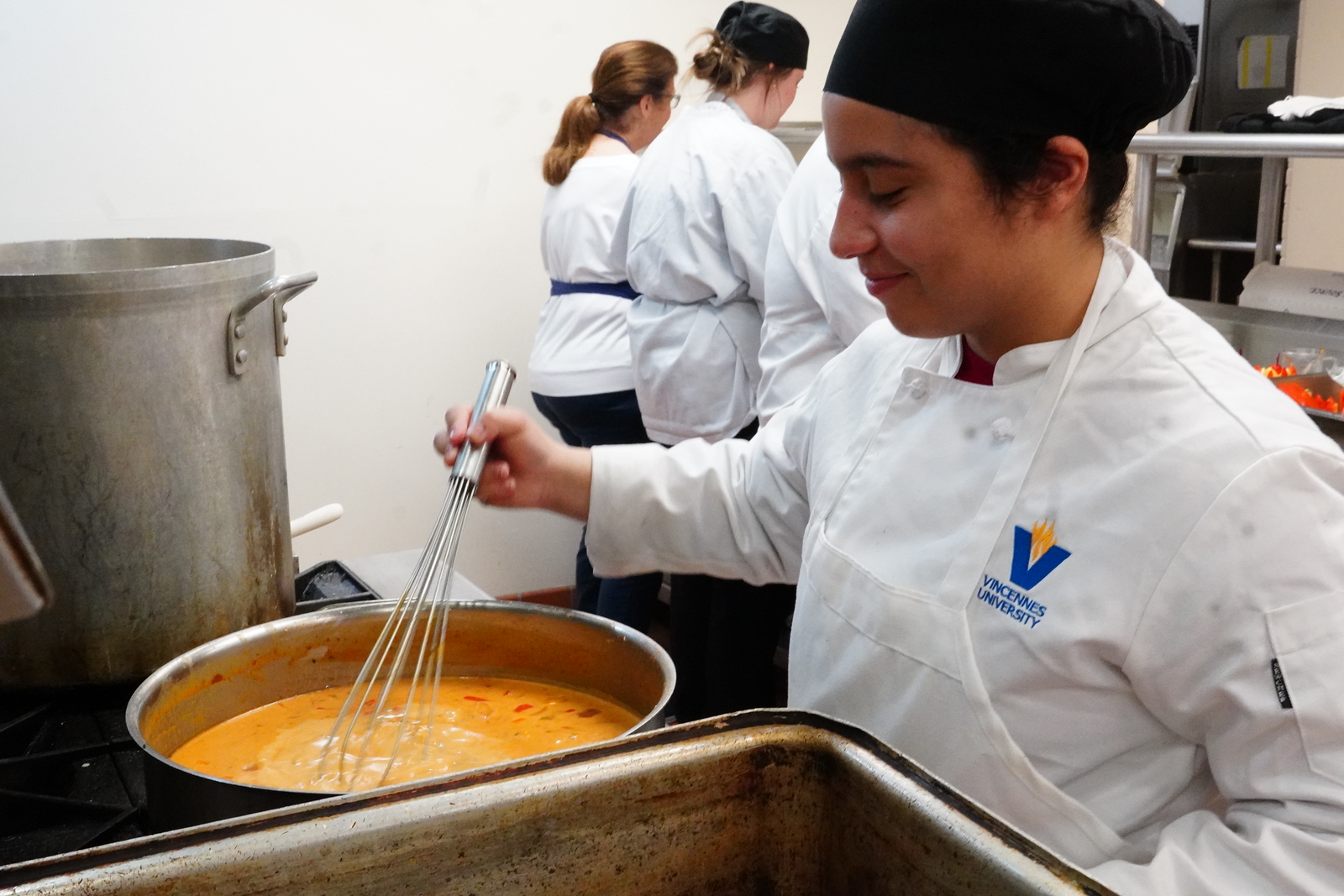 A Vincennes University culinary student stirs a large pot of soup while other students prepare other items for a meal.