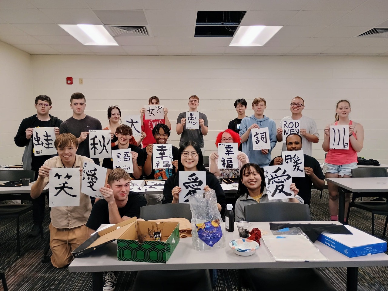 A classroom scene shows a group of approximately 15 students, many of whom are holding up sheets of paper displaying Japanese lettering.