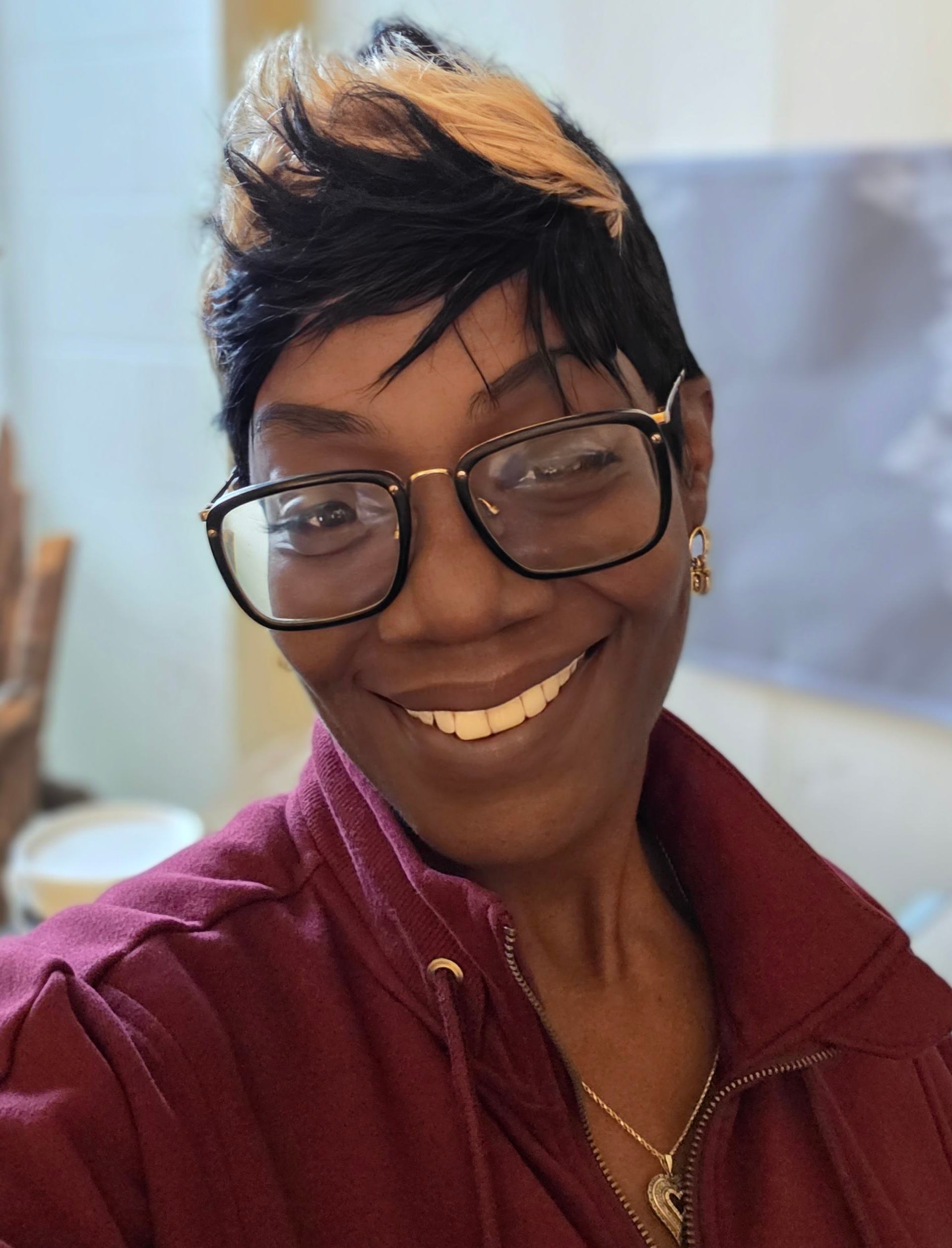 A smiling Lawana Dorsey with stylish short black and blonde hair wears glasses and a maroon zip-up jacket. She is indoors with a bright, warm expression.