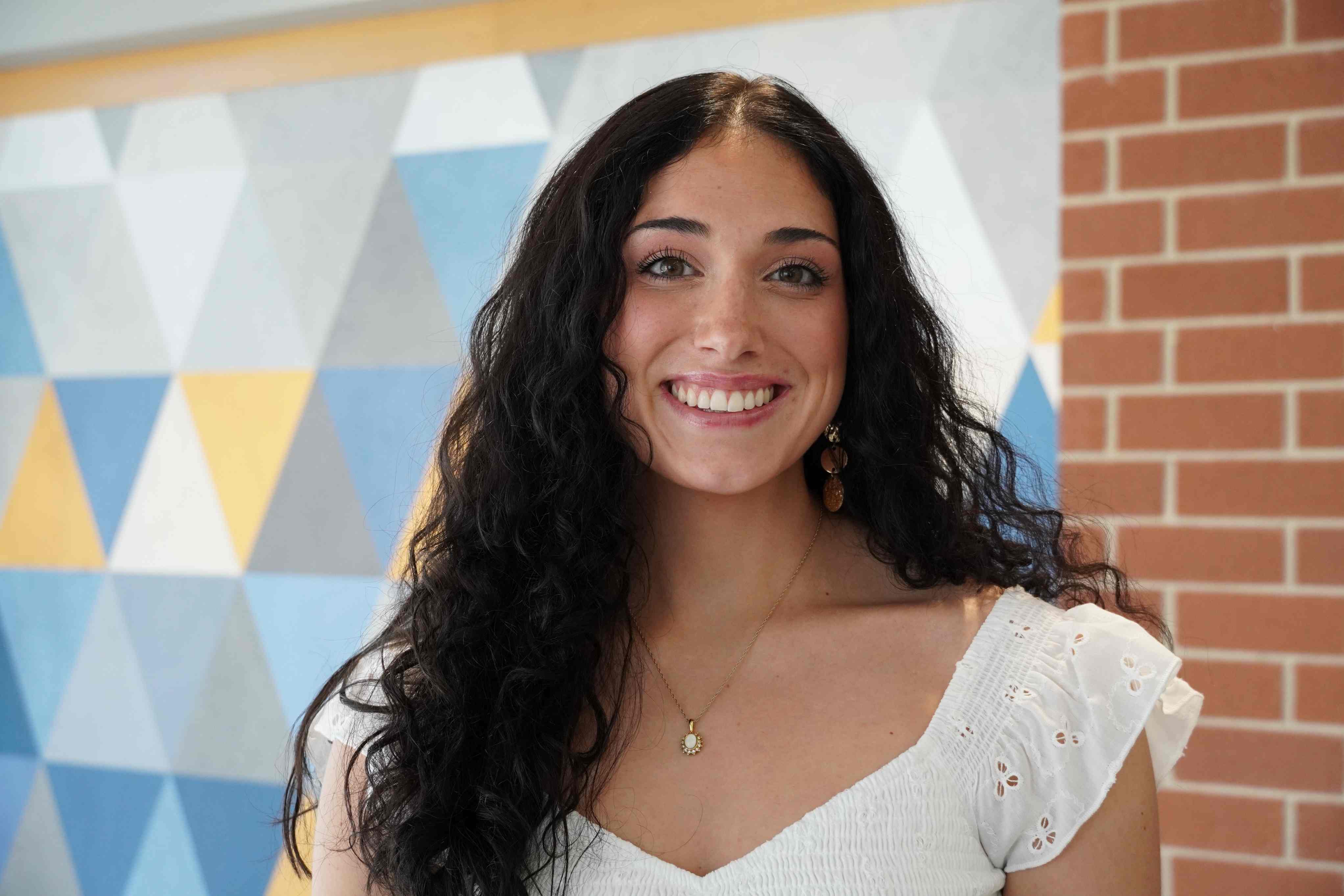  A smiling Maggie Lower with long, curly dark hair wearing a white ruffled top and gold necklace, posing against a yellow and blue geometric and brick wall background.