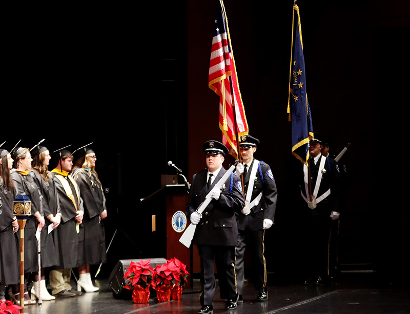 Graduation ceremony with a police honor guard carrying flags, standing on stage. Graduates in caps and gowns are lined up, and red poinsettias decorate the stage.