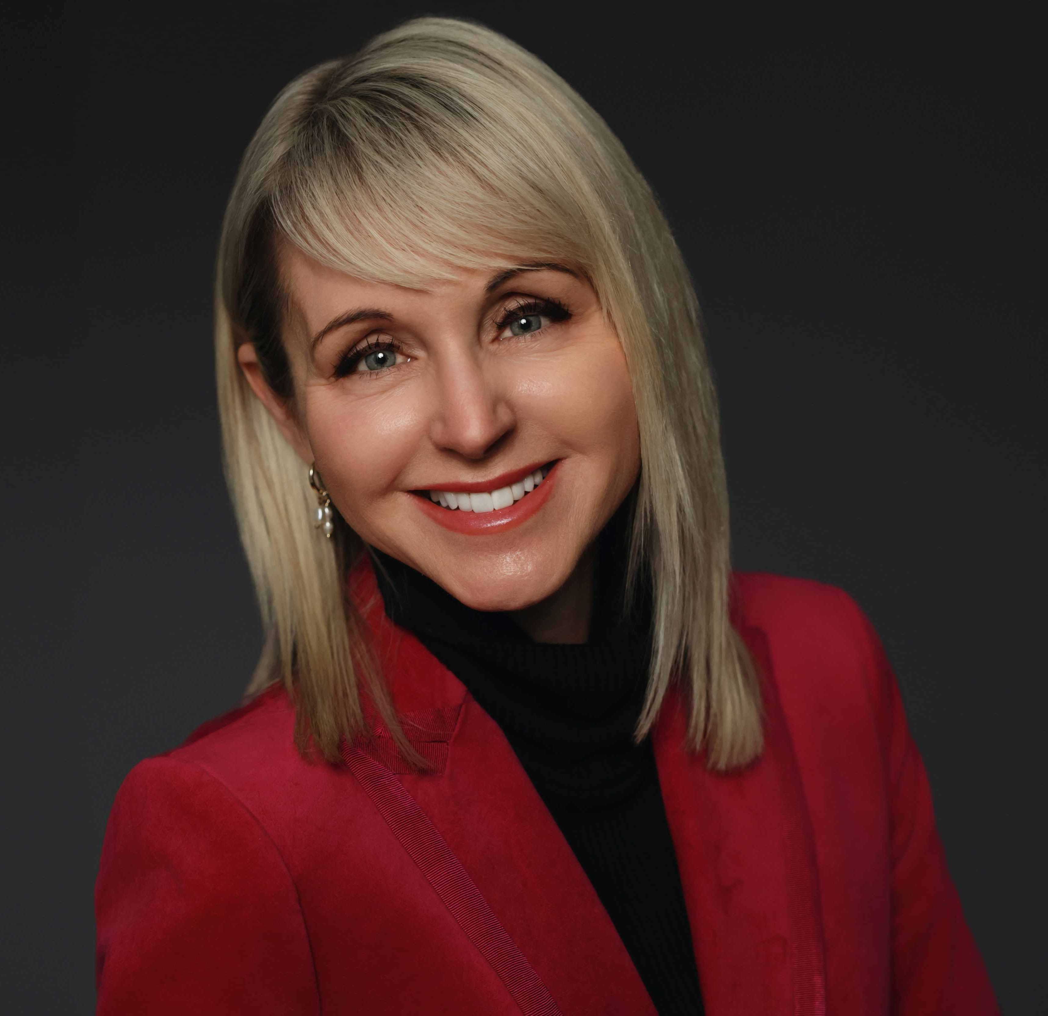 A professional headshot of Jaci Lederman with blonde hair, wearing a red blazer and black turtleneck against a dark gray background.