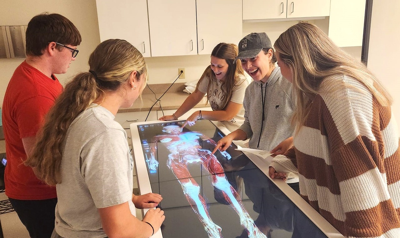 Four women stand at a large table-height monitor, looking intently at a brightly lit image of an androgynous human body with its skin and muscles rendered, with a white background.