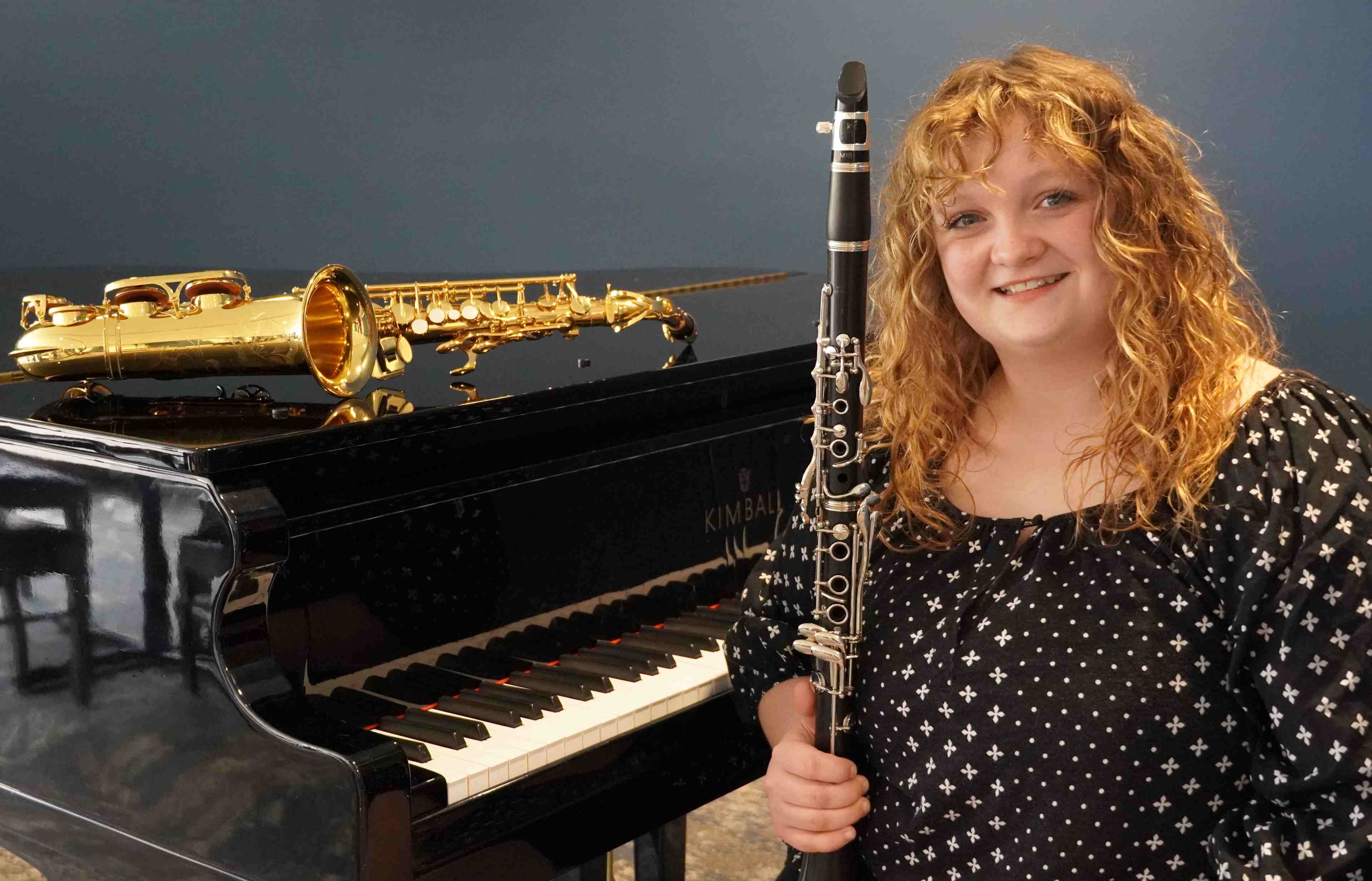 A smiling Savannah Wilson with curly hair holds a clarinet next to a black grand piano. A gold saxophone rests on top of the piano.