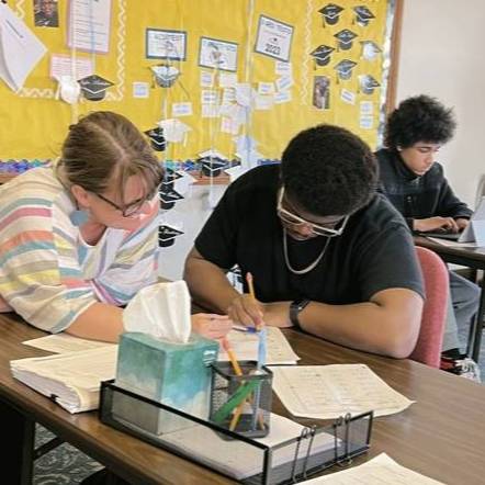 A student works with a teacher at their desk while another student works on an assignment in the background.
