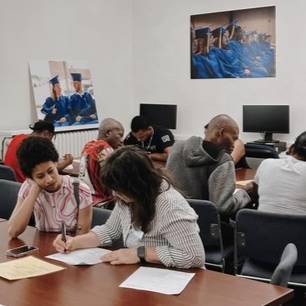 Two students work at a table while several other students work at computers behind them