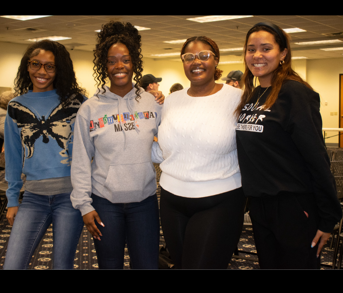 Four women stand together smiling in a room with rows of chairs. They're dressed casually in sweaters and hoodies, exuding a warm, friendly atmosphere.