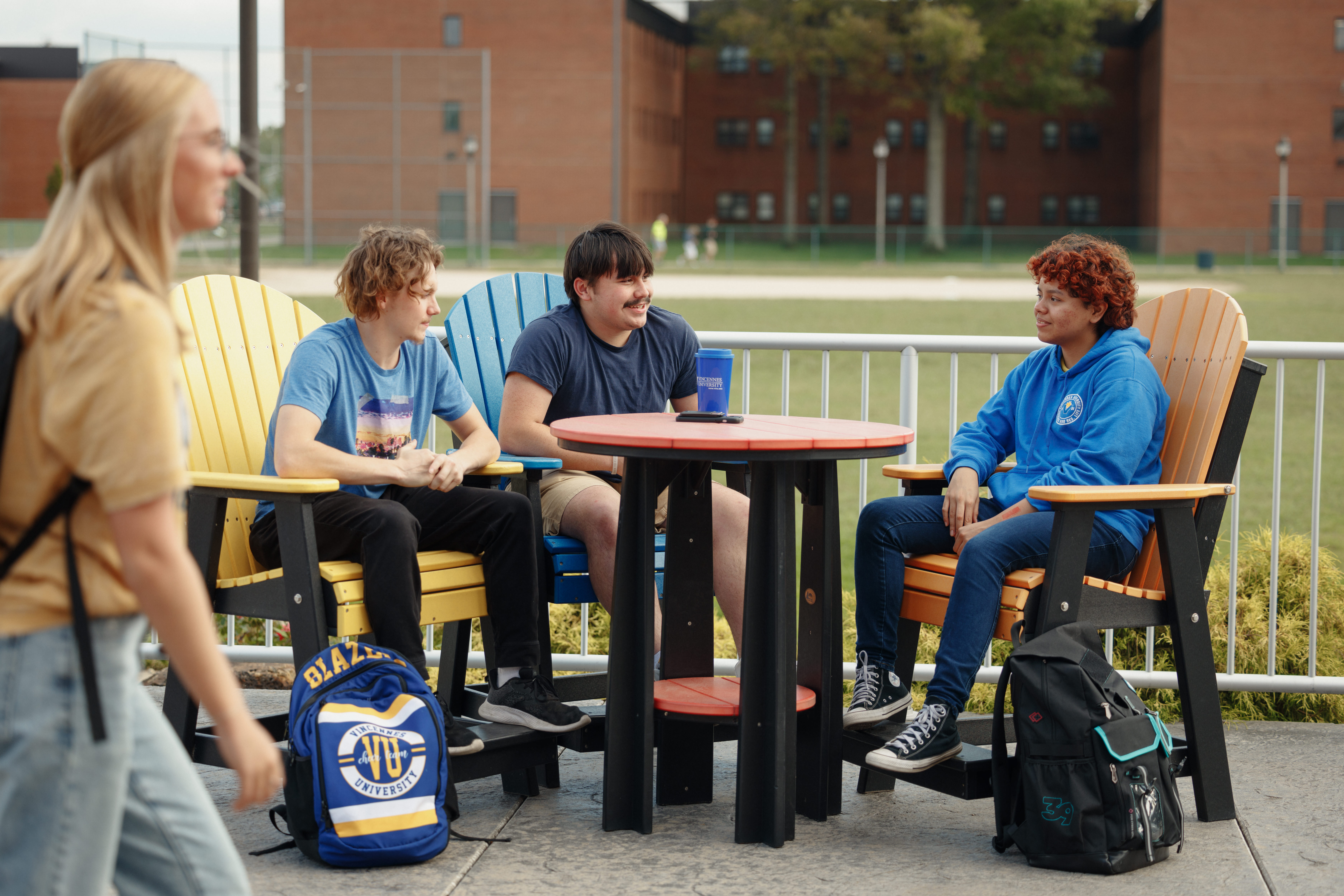 3 Vincennes University Students sitting outside chatting at a table with another student walking bu the group wearing a backpack