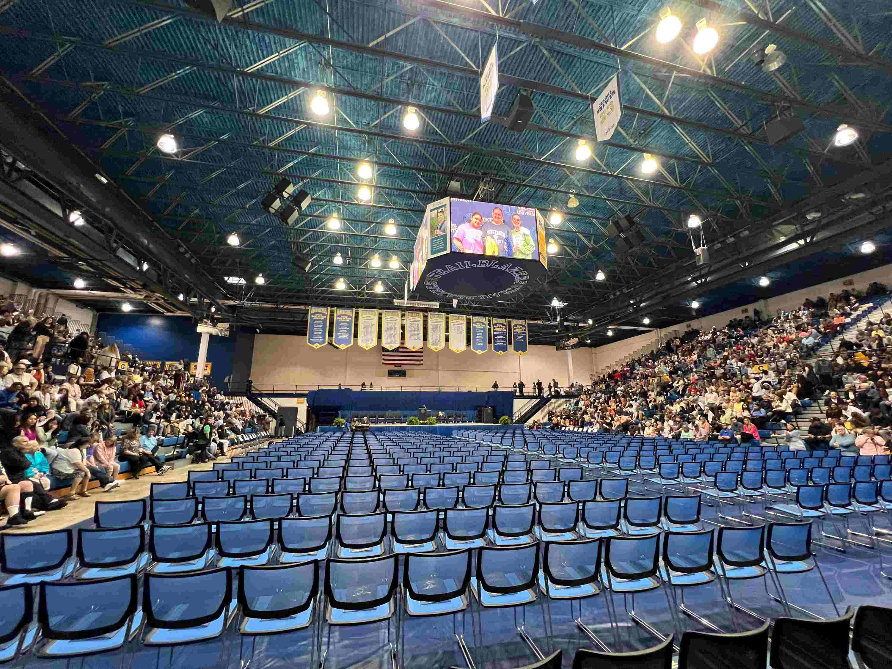 Interior view of a large, crowded VU Physical Complext set up for commencement with rows of blue chairs on the floor facing a stage and video board.
