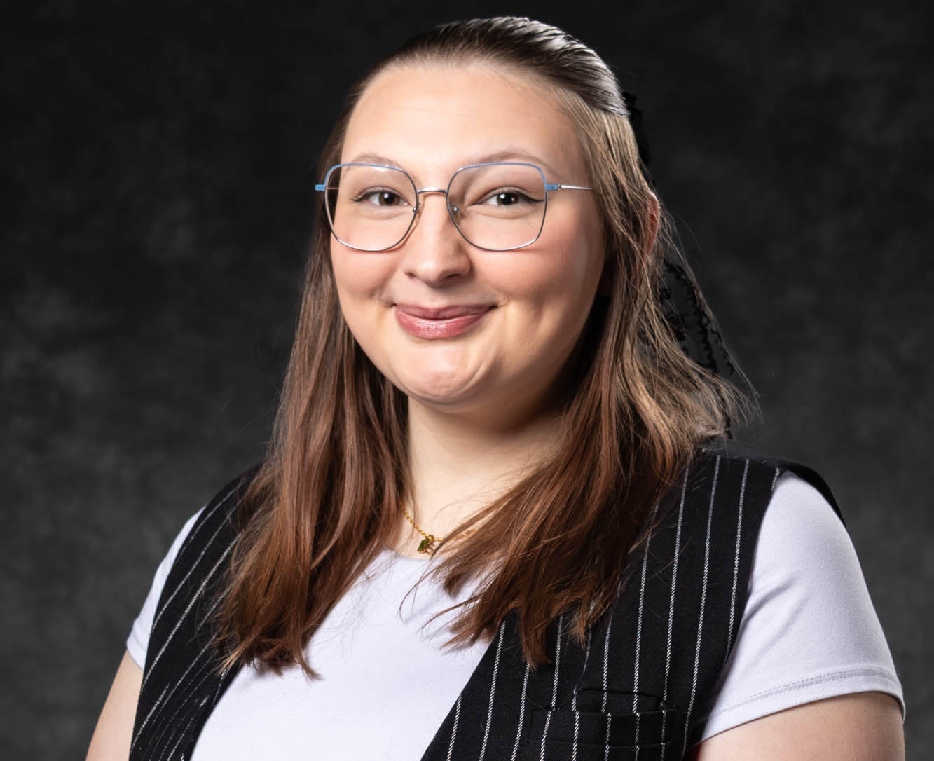 A smiling Zoey Rinesmith with glasses and brown hair, wearing a pinstripe vest over a white t-shirt, posed against a dark, textured studio background.